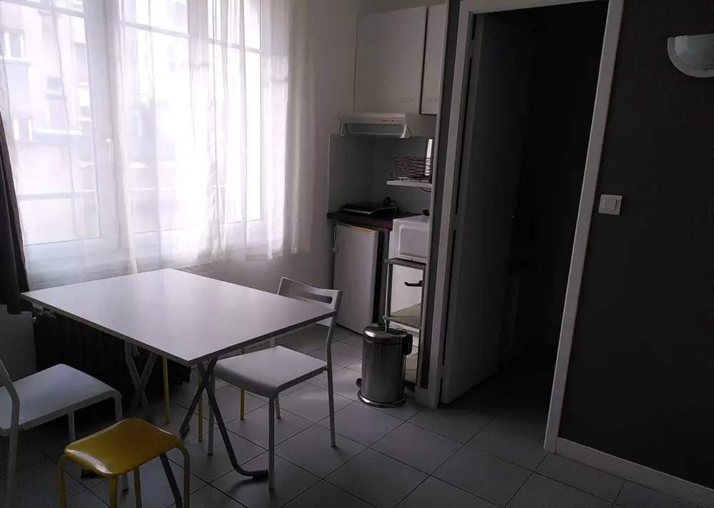 Le studio du 18 rue basse : coin repas près de la fenêtre, kitchenette et porte de la salle d'eau, sol carrelé clair.